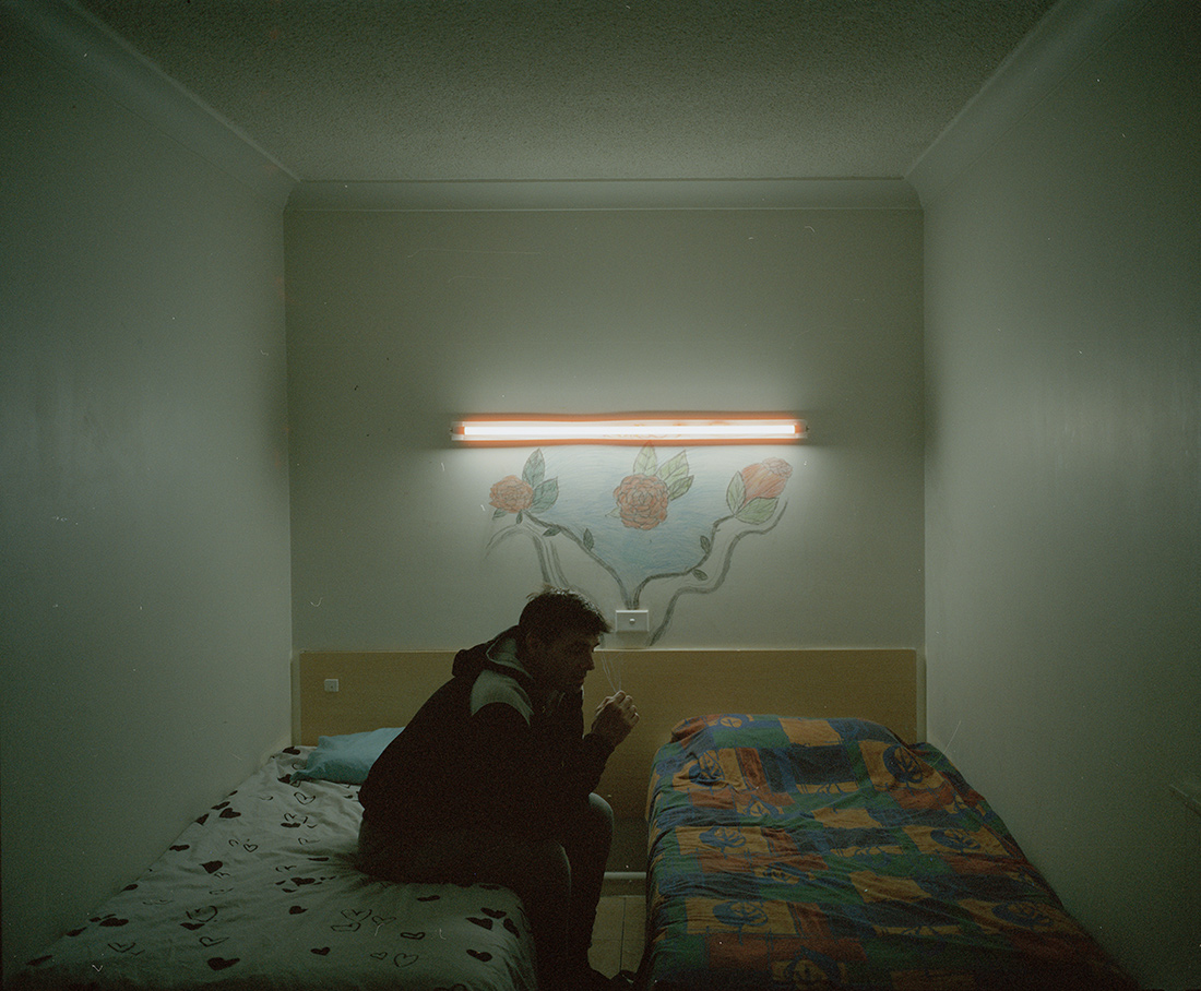 Adult male in a hooded jacket sits on one of two twin beds in an indoor setting with pale walls, a fluorescent light, and painted roses.