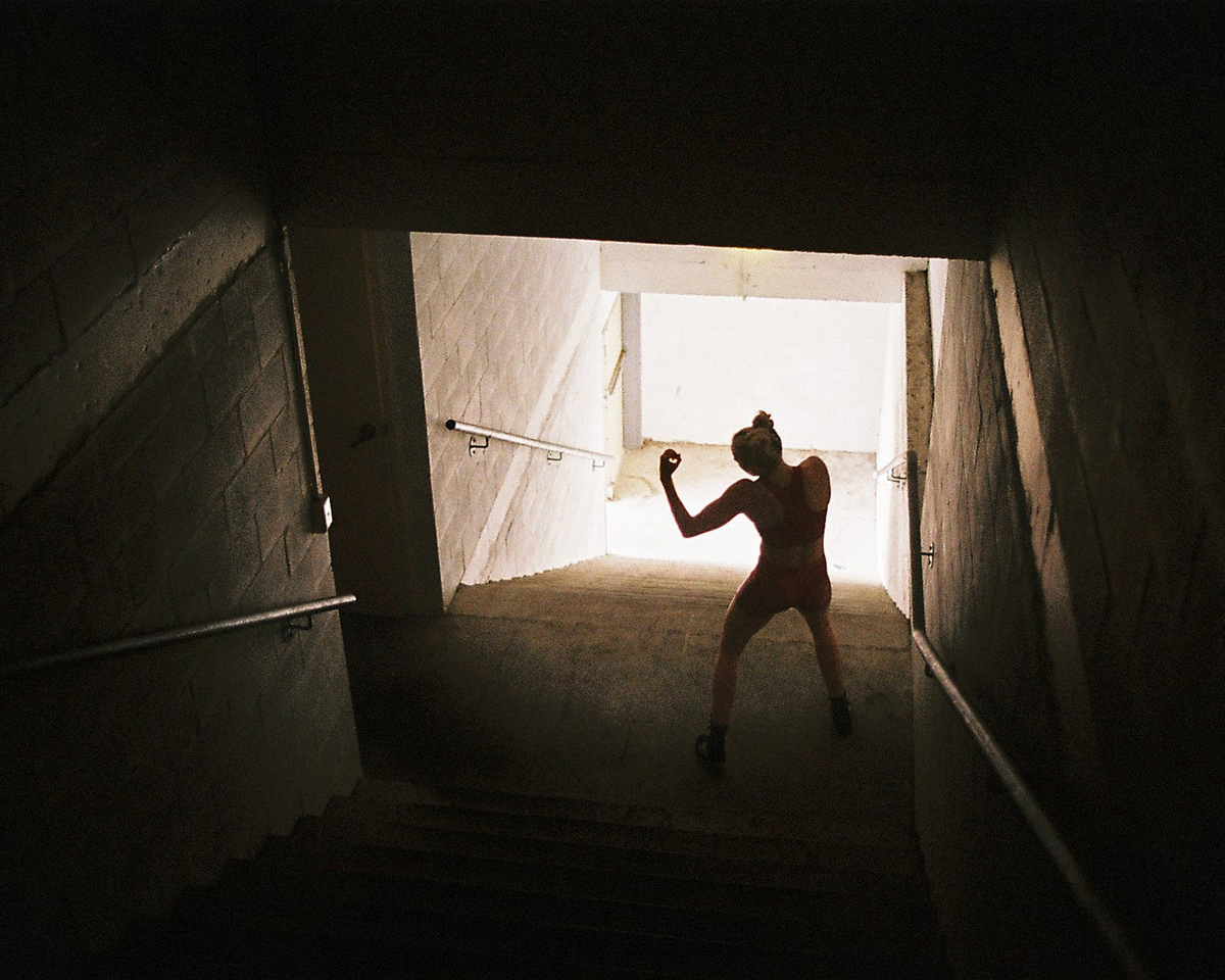 Woman in red athletic wear walks up dimly lit concrete steps towards a bright exit in a concrete tunnel setting. 