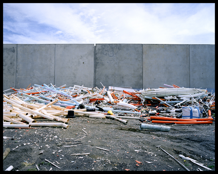 Outdoor shot of a pile of discarded PVC pipes and materials against a grey wall under a bright sky with scattered clouds.