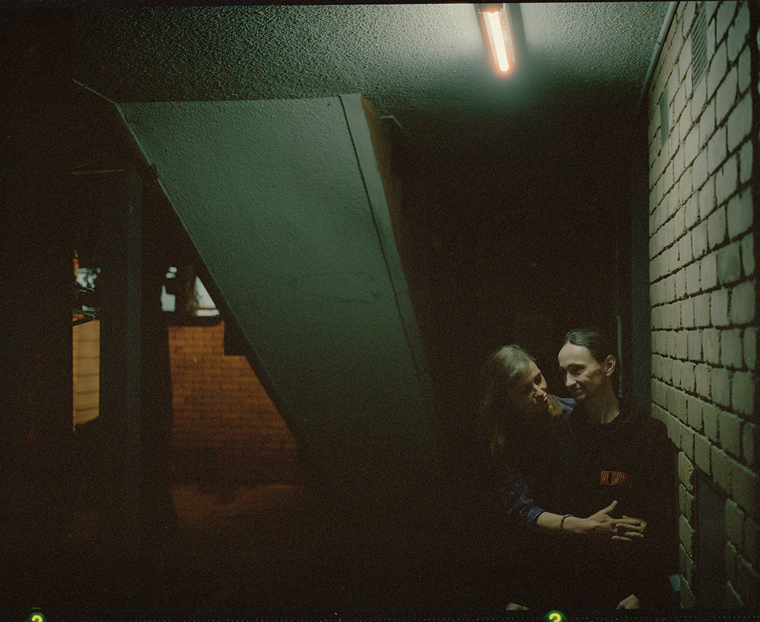 A couple embraces in the dim light of an urban stairwell with brick wall, concrete steps, and fluorescent lighting.