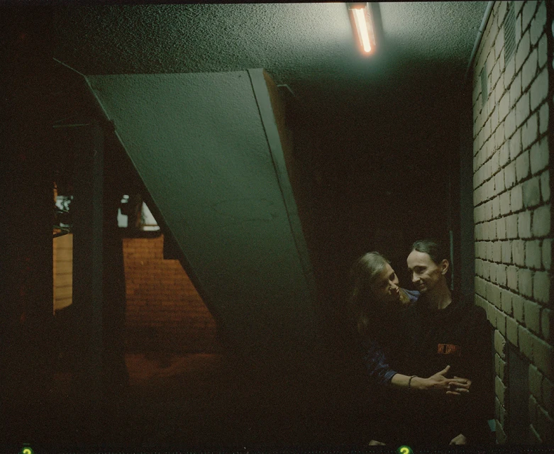 A couple embraces in the dim light of an urban stairwell with brick wall, concrete steps, and fluorescent lighting.