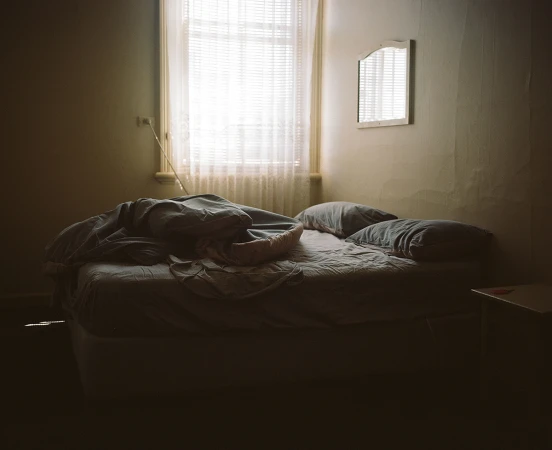 Unmade bed in a dimly lit bedroom with sunlight streaming through a lace-curtained window.