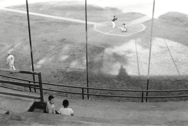 High angle black and white view of two children sitting in empty stadium bleachers watching a baseball game through a chain link fence.