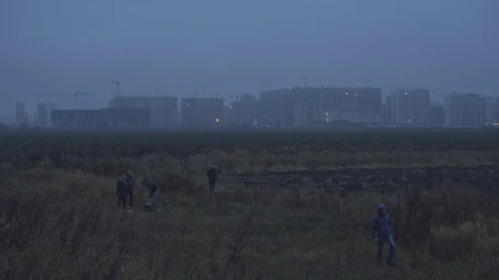 Distant figures standing in a vast, dark field at dusk with silhouetted high-rise buildings and construction cranes visible in the background