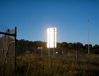 A reflective sign glows brightly in front of a chain-link fence and tall grass under a clear blue sky, captured by Sherwin Akbarzadeh.