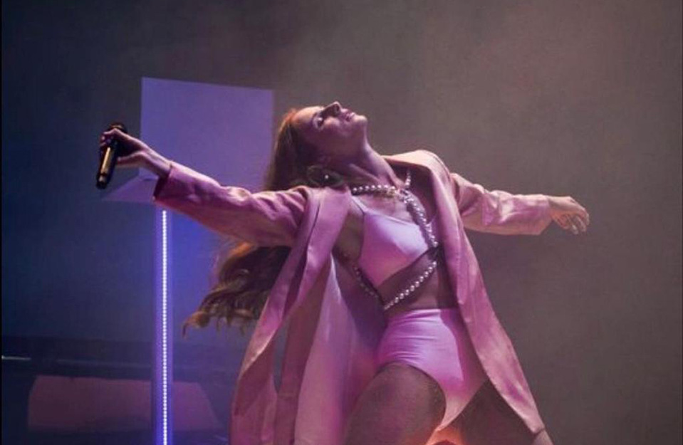 A woman in a pink outfit and coat, wearing a pearl body chain, stands on a stage with a microphone, singing and dancing during a performance.