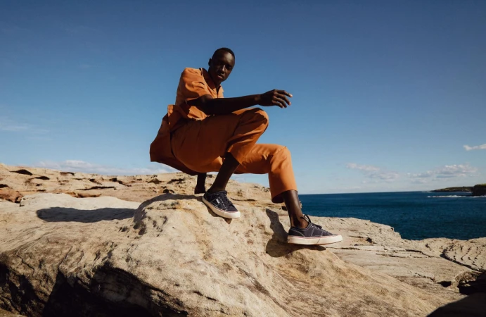 Black man crouching on rocky coastal ledge wearing rust orange shirt and pants with blue sneakers against ocean backdrop