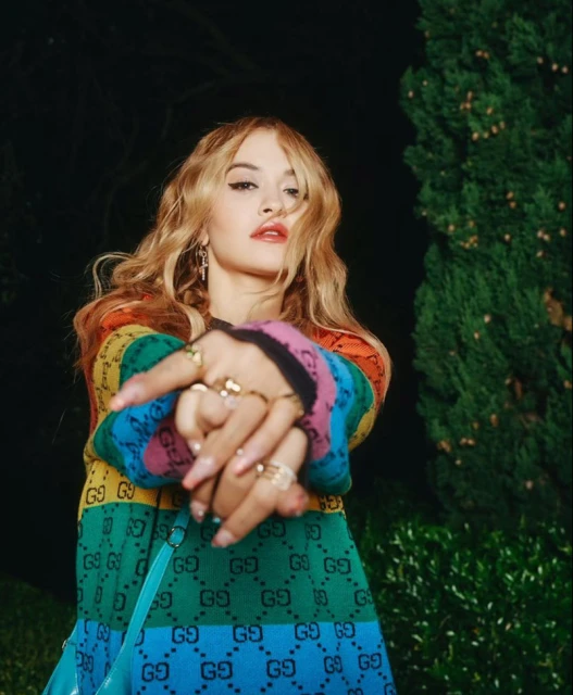 Portrait of a stylish young woman with blond hair, wearing a colorful sweater and jewelry, posing outdoors at night with greenery in the background.