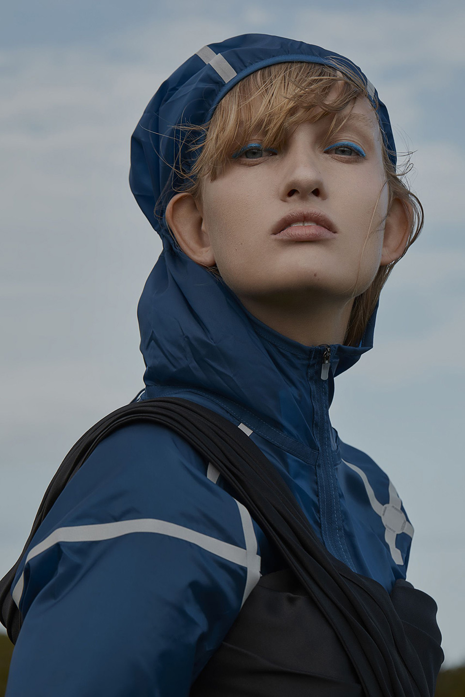 Fashion portrait of a young blonde model with blue eyeshadow, wearing a hooded jacket and a black top, set against a cloudy sky.