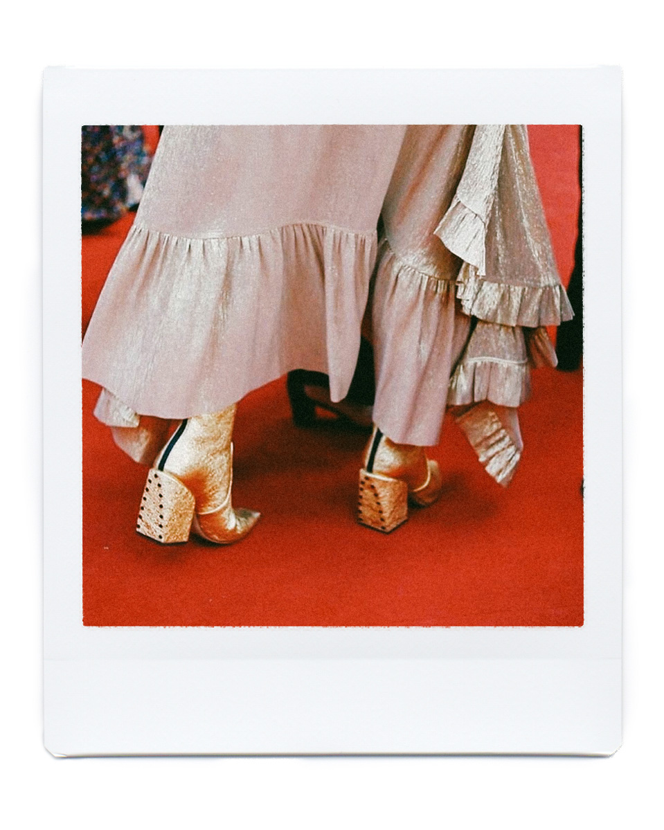 Close-up of gold, block-heeled ankle boots on a red carpet, paired with a ruffled champagne skirt, in a Polaroid-style frame.
