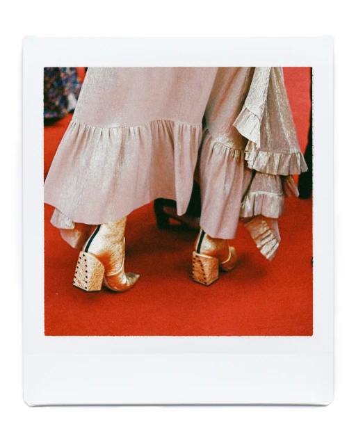 Close-up of gold, block-heeled ankle boots on a red carpet, paired with a ruffled champagne skirt, in a Polaroid-style frame.