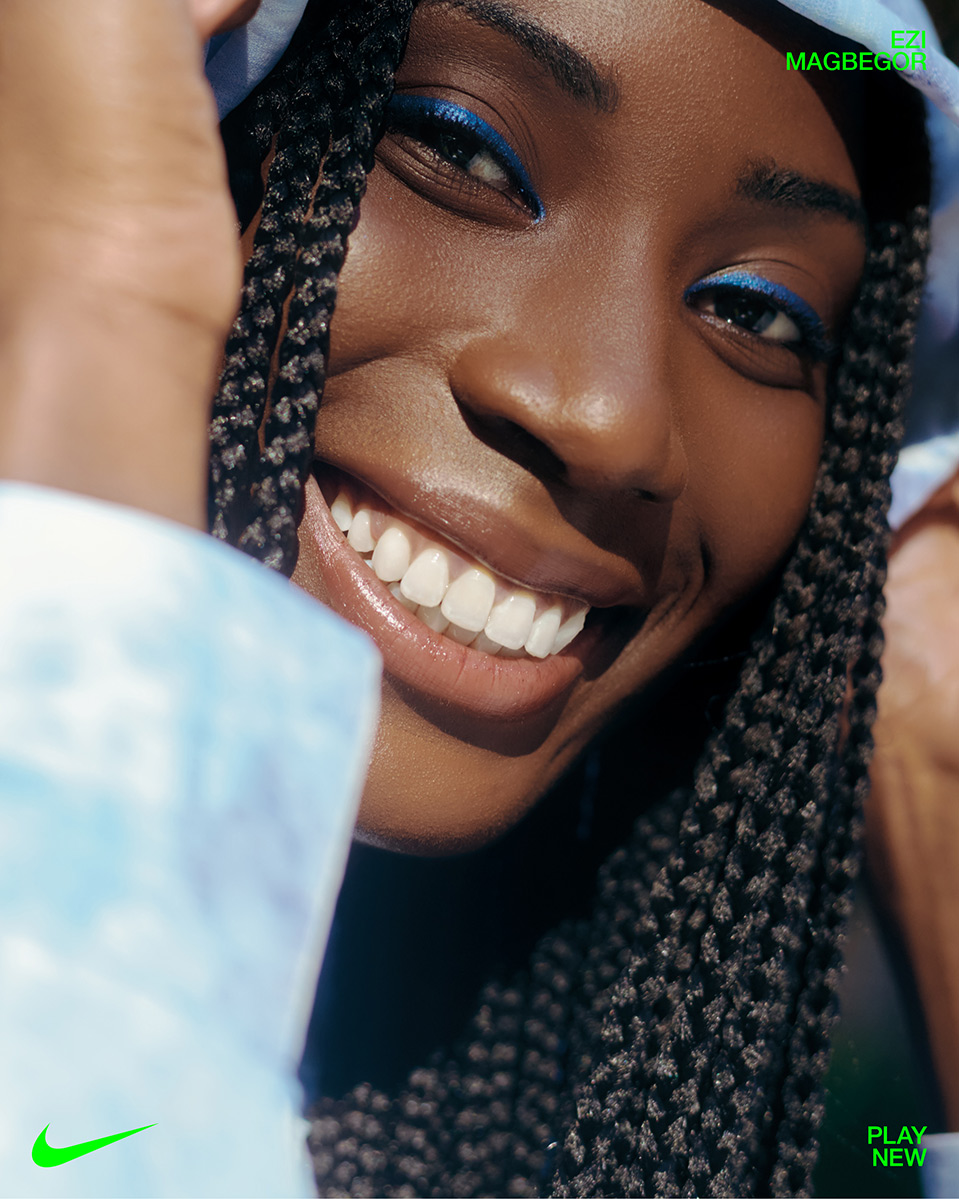 Close-up of a smiling woman with braided hair, bright blue eyeliner, and a light blue headscarf, with a Nike logo in the corner.