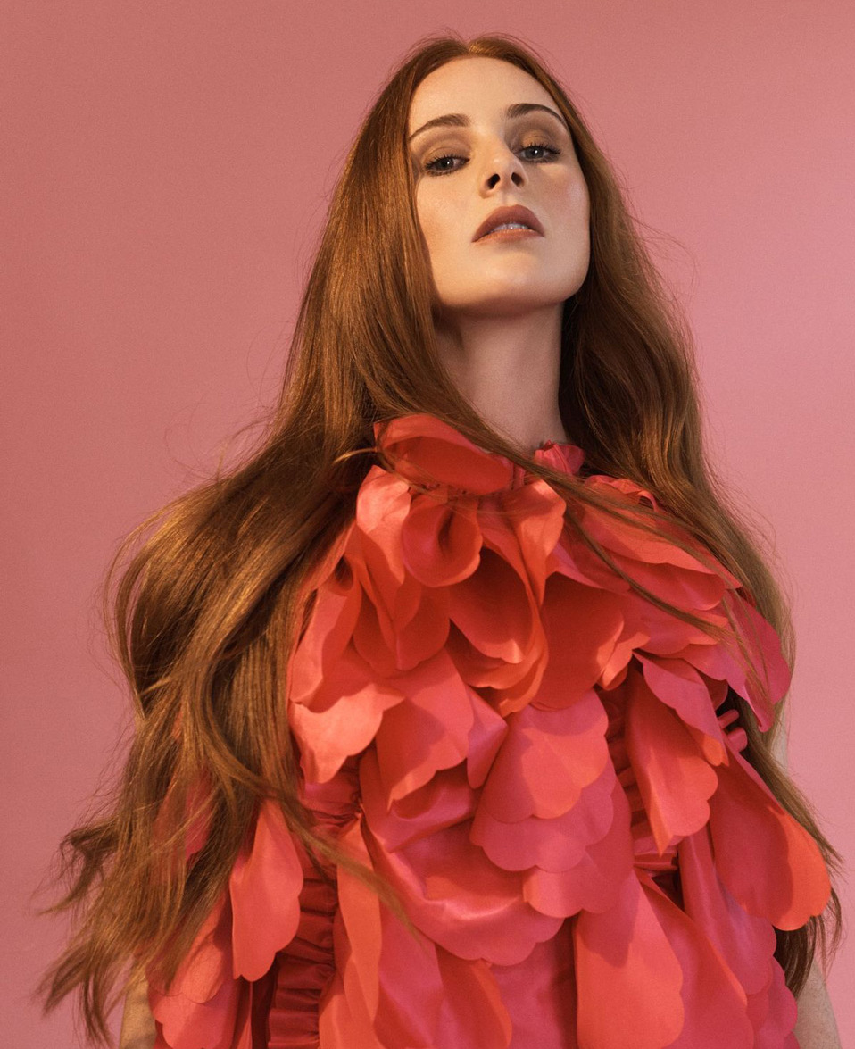 Close-up portrait of a woman with long red hair posing in a pink, floral dress against a pink backdrop in a photo studio.
