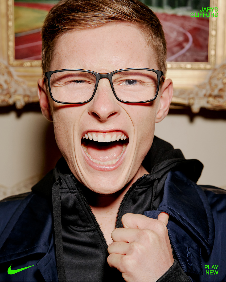 Close-up portrait of a Caucasian adult male athlete with glasses and an open mouth, wearing a navy tracksuit jacket indoors.
