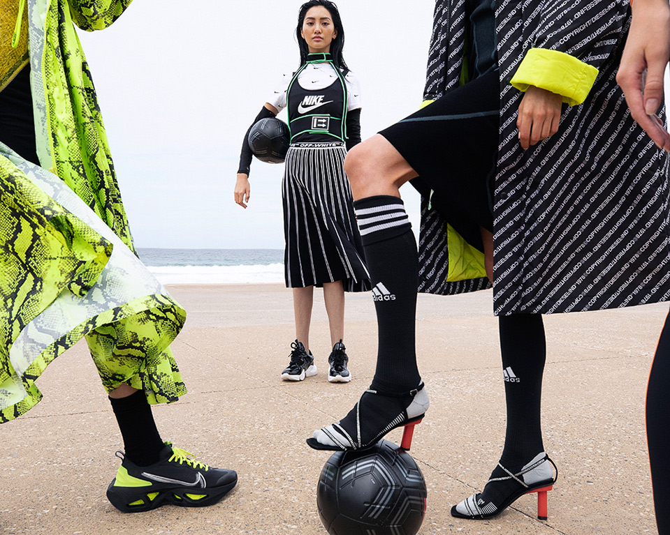 Three women on a beach in designer sport clothing featuring Nike and Adidas brands with a soccer ball. Editorial fashion shoot.