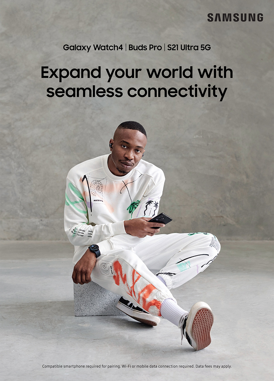 Samsung ad featuring an adult male wearing ear buds, a smartwatch, and holding a smartphone while sitting against a gray background.