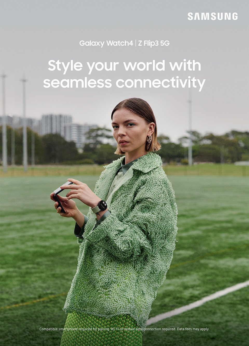 A woman in a green textured jacket and skirt stands on a green field holding a smartphone, wearing a smartwatch in a Samsung advertisement.