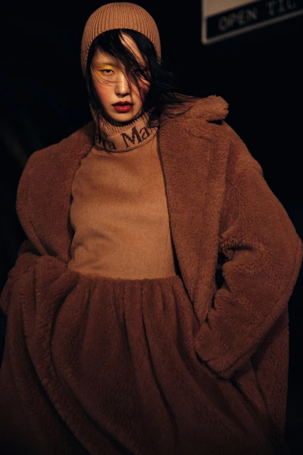 Asian woman poses in a brown teddy coat, turtleneck, and matching beanie, set against a dark background with partial signage visible.
