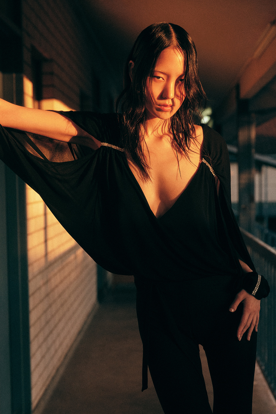 Asian woman in a black dress poses indoors with sunlight casting shadows on the wall. Fashion editorial, portrait, full body.