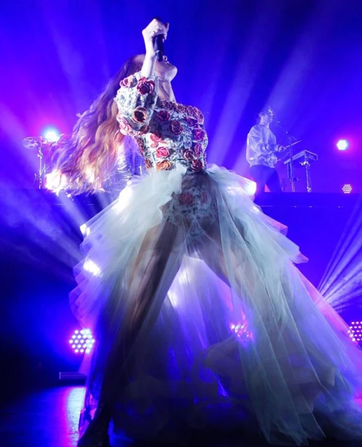 Female musician in floral dress performs on stage with microphone, backed by band member in concert lighting; long hair flows with energy.