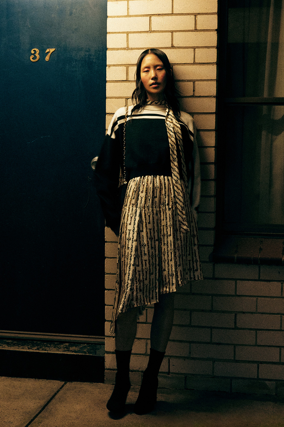 Young Asian woman in patterned dress poses against brick wall next to blue door with '37', soft directional lighting, dark ankle boots.
