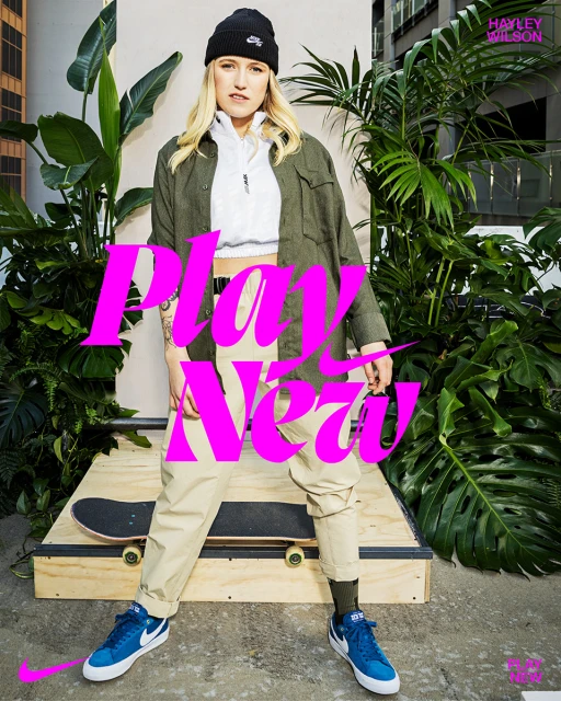 Young woman posing with a skateboard, wearing a beanie, jacket, and sneakers, standing outdoors with plants and an urban backdrop.