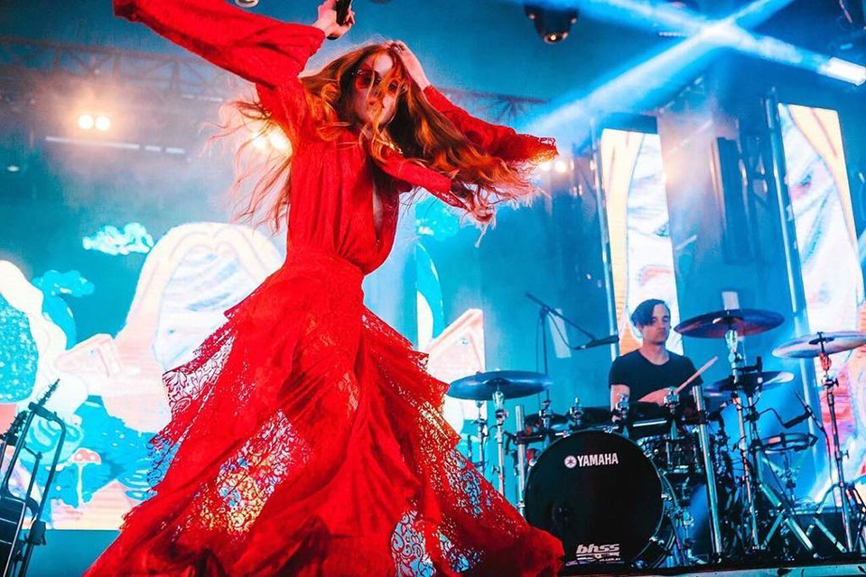 A woman wearing a red dress and holding a microphone performs on a stage with a drummer and musical equipment in the background.