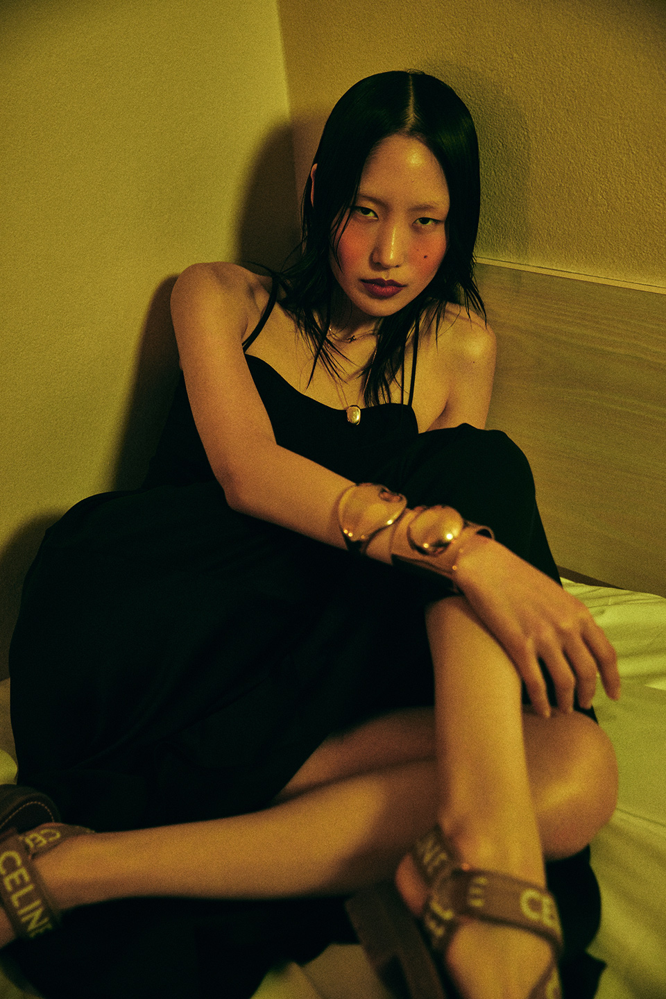 An Asian woman sits indoors wearing a black dress, sandals, and gold bangles. She poses against a yellowish wall with neutral lighting.