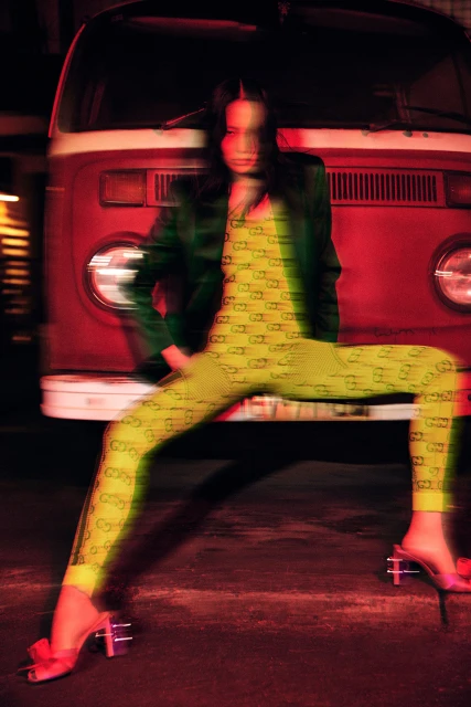 A woman in a patterned outfit poses in front of a red VW bus, shot with motion blur and low lighting for a dynamic fashion image.