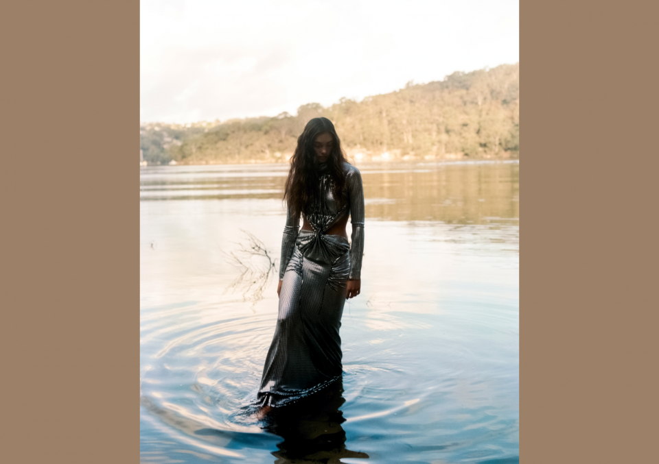 An adult woman stands knee-deep in a lake, wearing a shimmery silver gown with a cut-out bodice, under soft natural light and trees in the background.