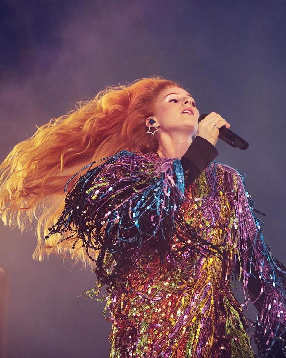 An adult female singer with red hair holds a microphone while singing on stage in a fringed sequined dress, with dramatic lighting.