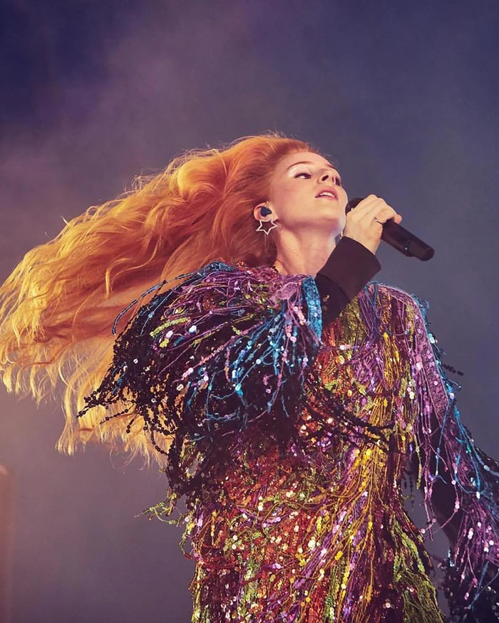 Female singer with long blowing red hair wearing a colourful sequin fringed jacket singing into a microphone on stage.