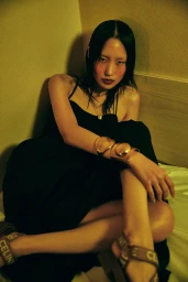 Asian woman in a black dress with gold bracelets and sandals seated on a bed styled by Jana Bartolo.