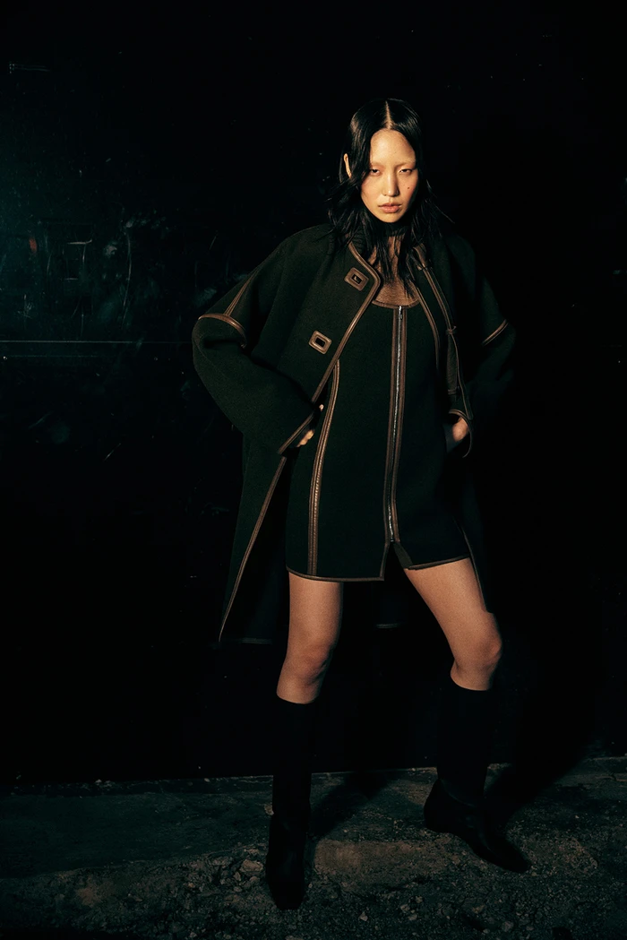 Model wearing dark coat and mini dress with contrast trim, styled by Jana Bartolo against a dark textured background