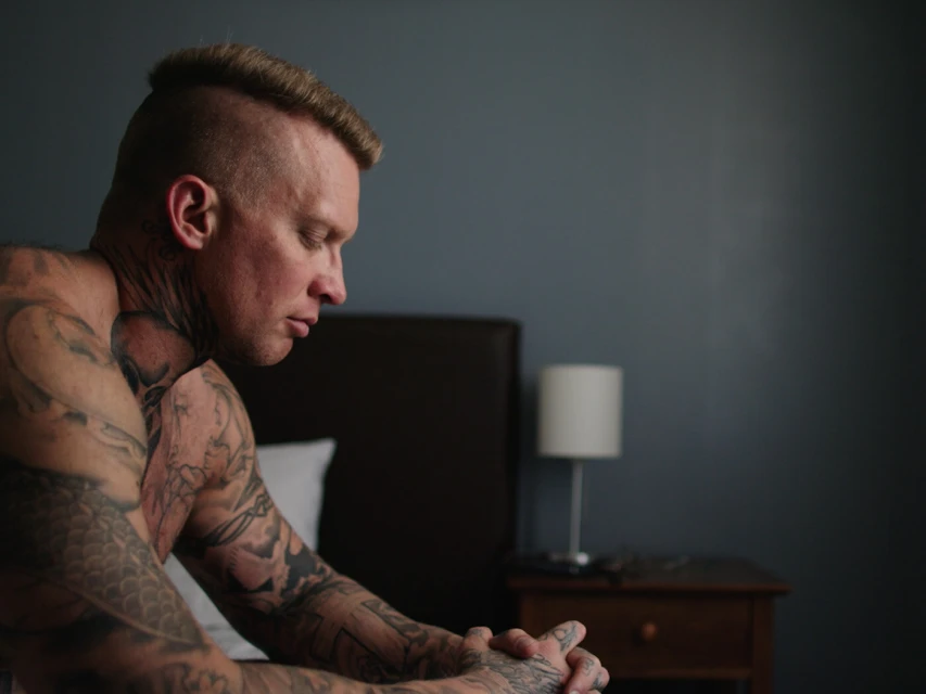Adult male with tattoos sits indoors on a bed in contemplation with hands clasped. Soft natural light from the side illuminates the scene.