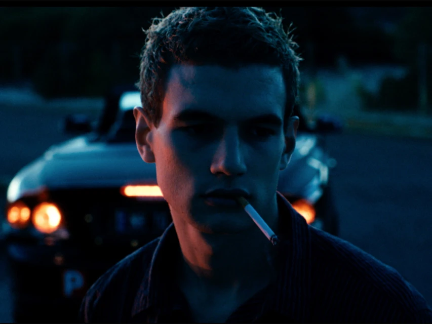 Adult male with cigarette in his mouth stands outdoors at night in front of a car with headlights and a dark blue background.