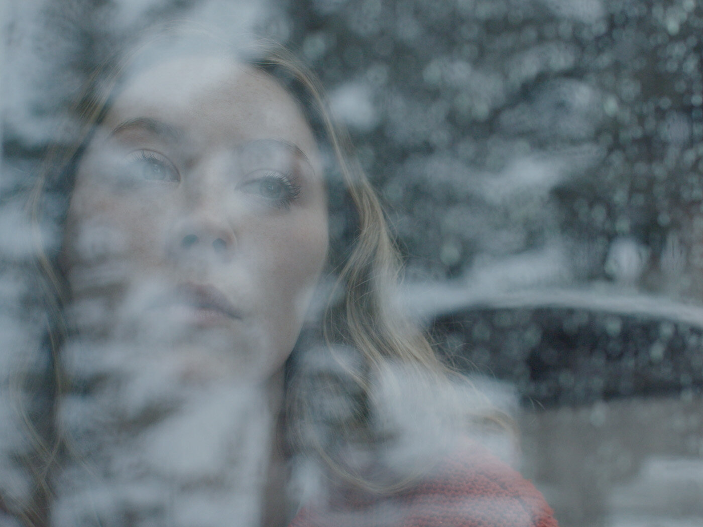 Close-up portrait of a woman looking through a rain-streaked window with soft, desaturated lighting and visible reflections.