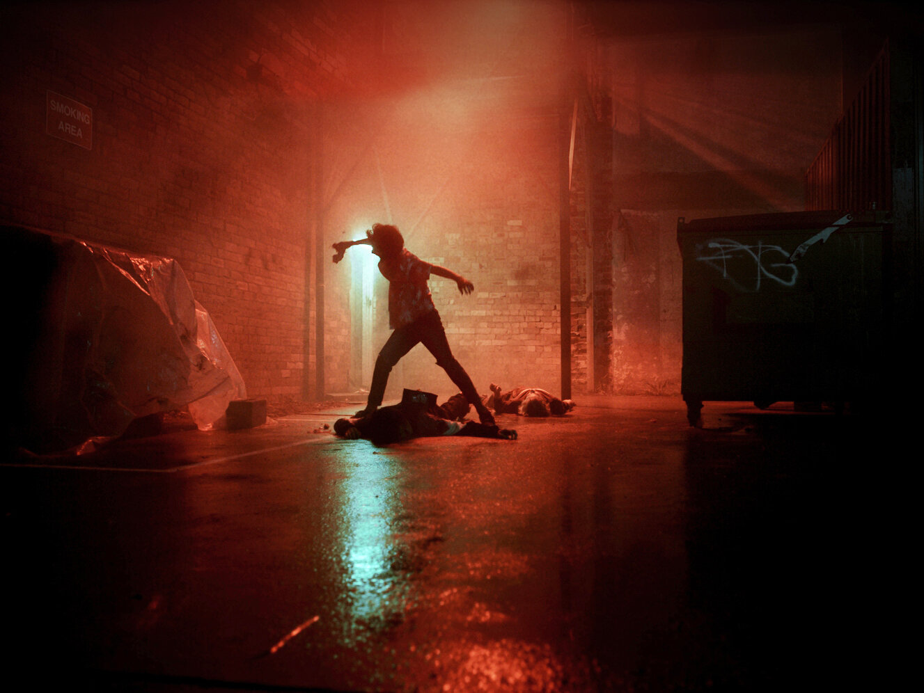 A man stands over two prone figures in an urban alleyway, illuminated by red and cyan light, creating a dramatic, edgy atmosphere.