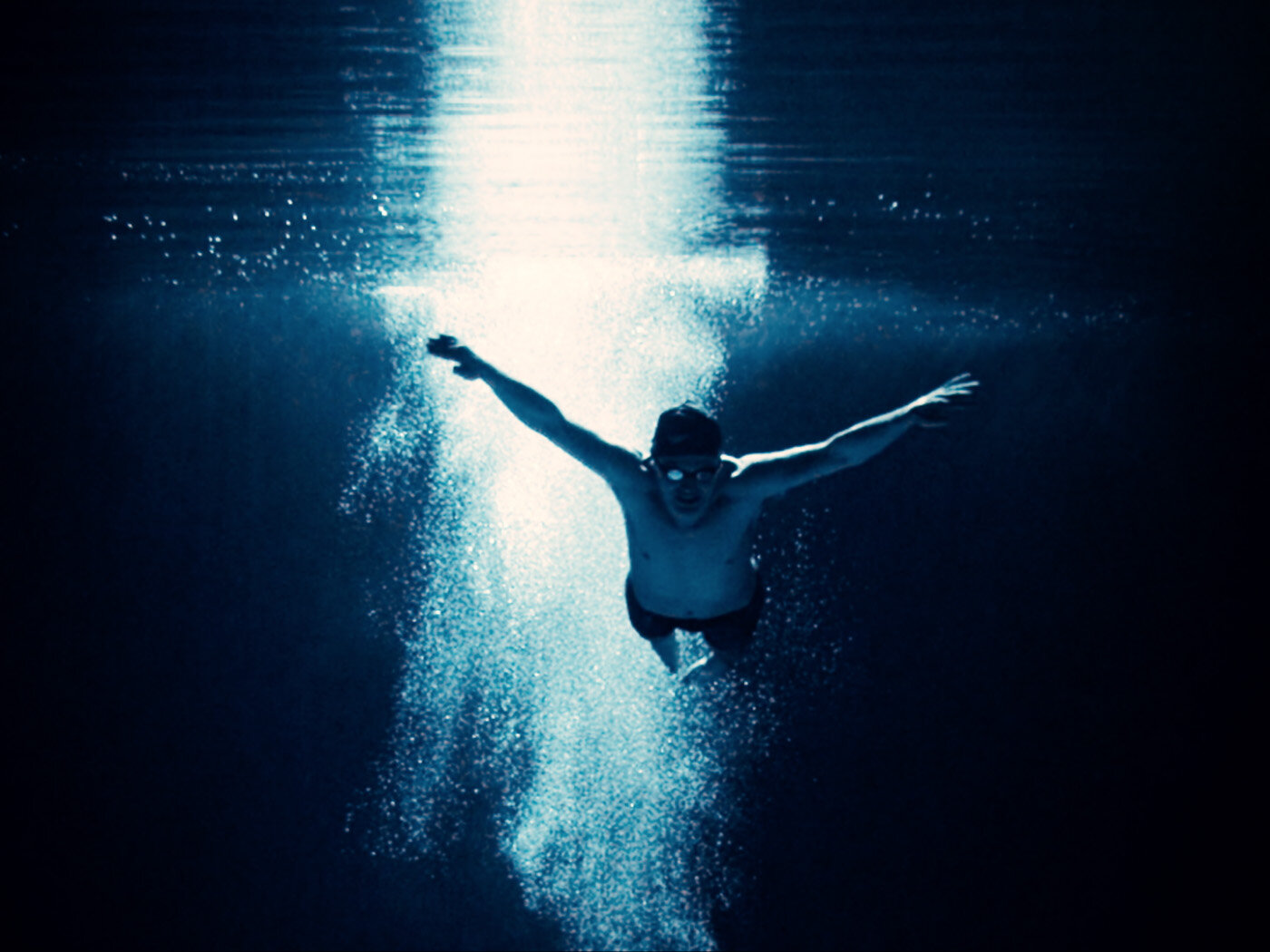 Underwater shot of a swimmer with arms outstretched, wearing goggles, surrounded by bubbles, against a bright light in deep blue water.