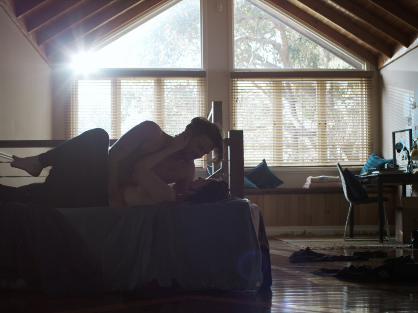 Two people laying together on a bed inside a sunlit room. The composition is intimate, highlighting the connection between the subjects.