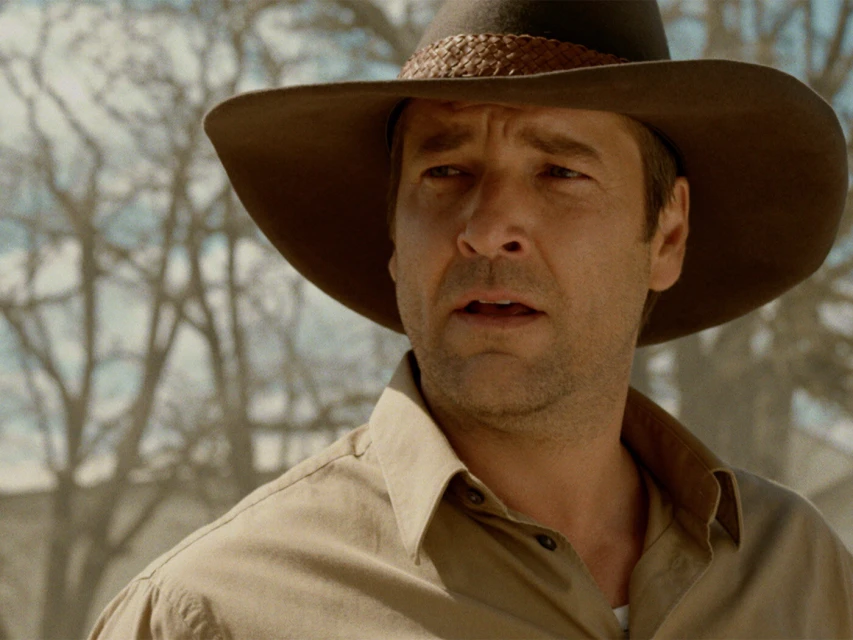 Close-up of an adult male wearing a tan hat and shirt outdoors in a bright and sunny environment with bare trees in the background.