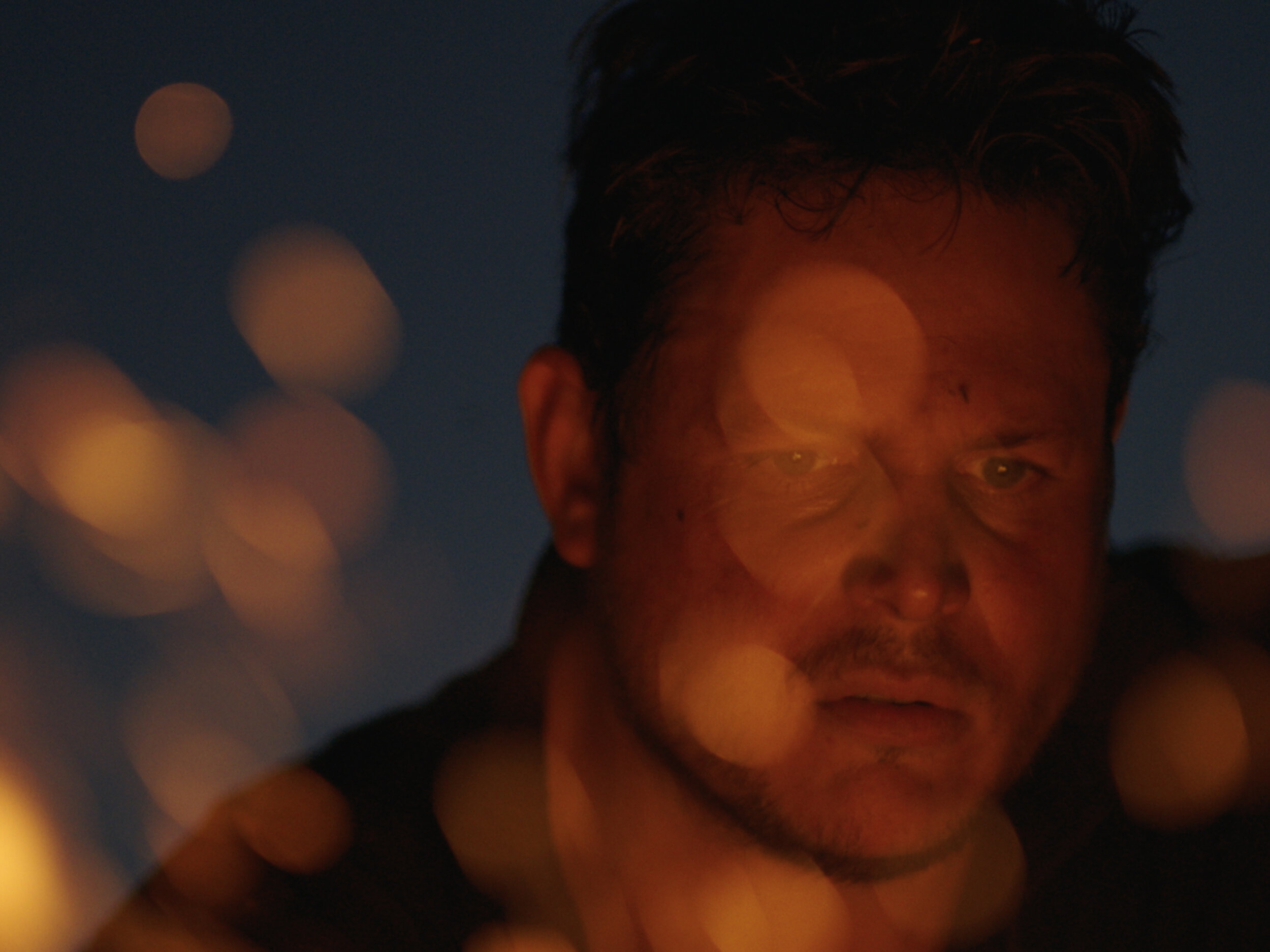 Close-up of a man's face illuminated by orange bokeh lights against a dark blue background, creating a dramatic, intimate portrait.