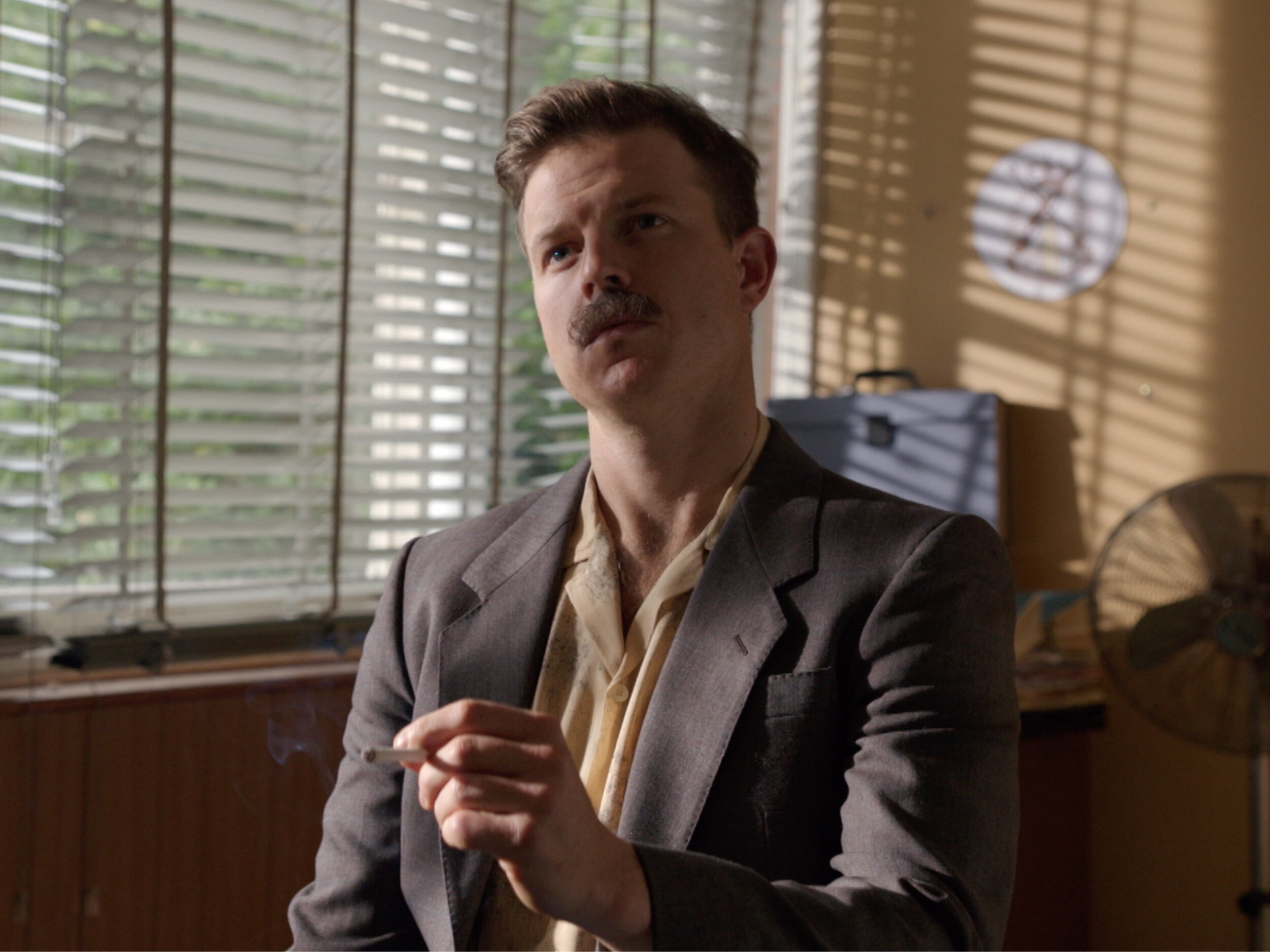 Portrait of a man with a mustache holding a cigarette indoors with light shining through window blinds and a fan in the background.