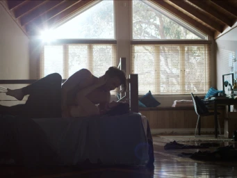 A man and a woman embracing intimately on a bed in a sunlit bedroom with large wooden-framed windows and vaulted ceilings.