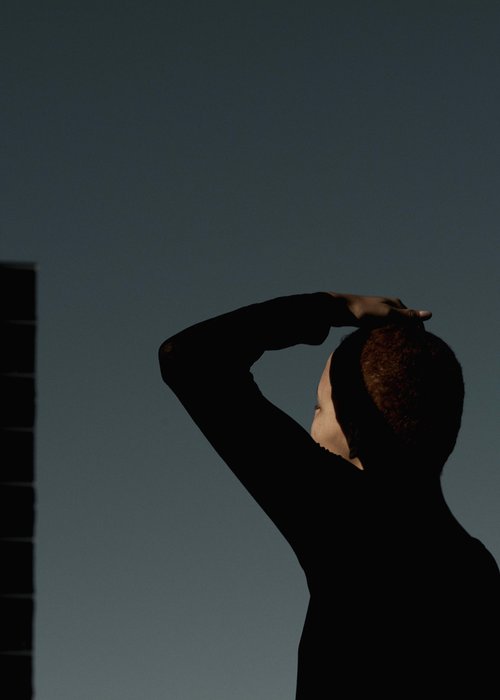 Silhouetted figure with red hair stands against a blue sky, partially obscured by the shadow of a building on the left.