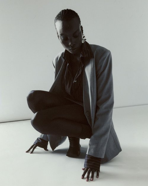 A young woman with dark skin poses in a grey blazer and black shirt against a bright white background, squatting and looking down.