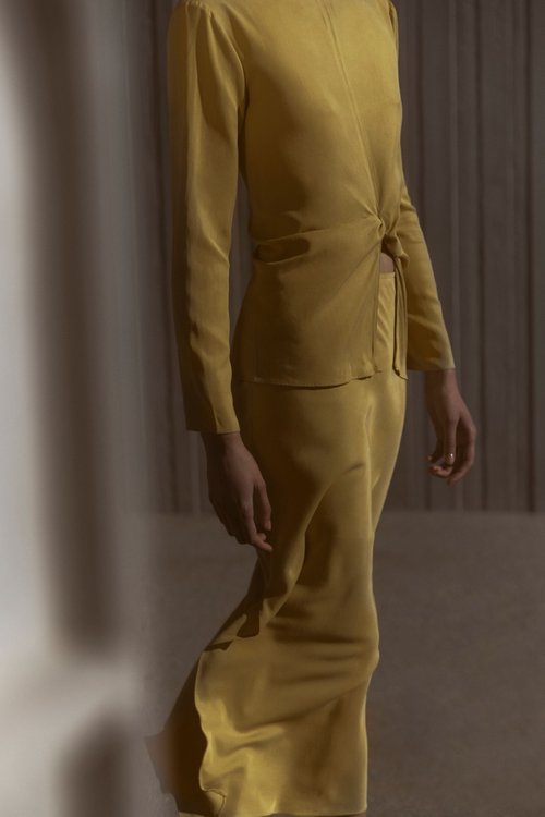 Woman in a golden yellow long sleeve blouse and skirt with a knot detail, standing in an indoor setting with natural lighting.