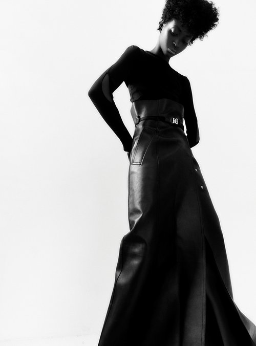 Black model in a long-sleeved black top and a long black leather skirt posing against a white background in a fashion shot.
