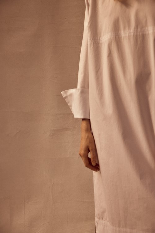 Close-up of a person wearing a white oversized shirt with long sleeves, posing against a textured, light brown wall in soft lighting.
