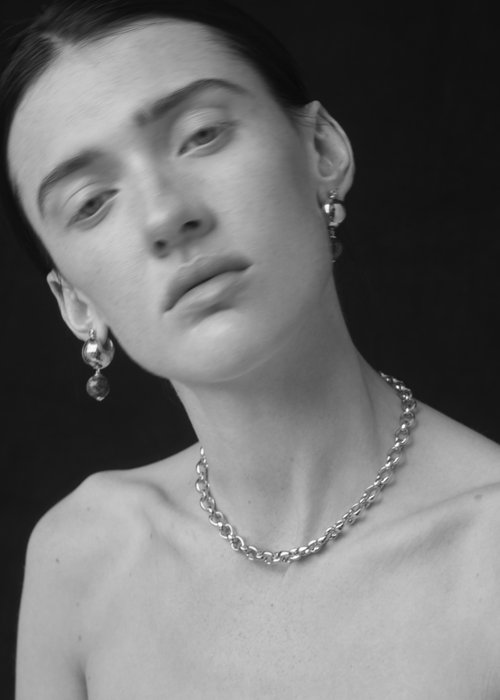 Monochrome portrait of a young adult woman with bare shoulders, wearing a chain necklace and round earrings, against a dark background.